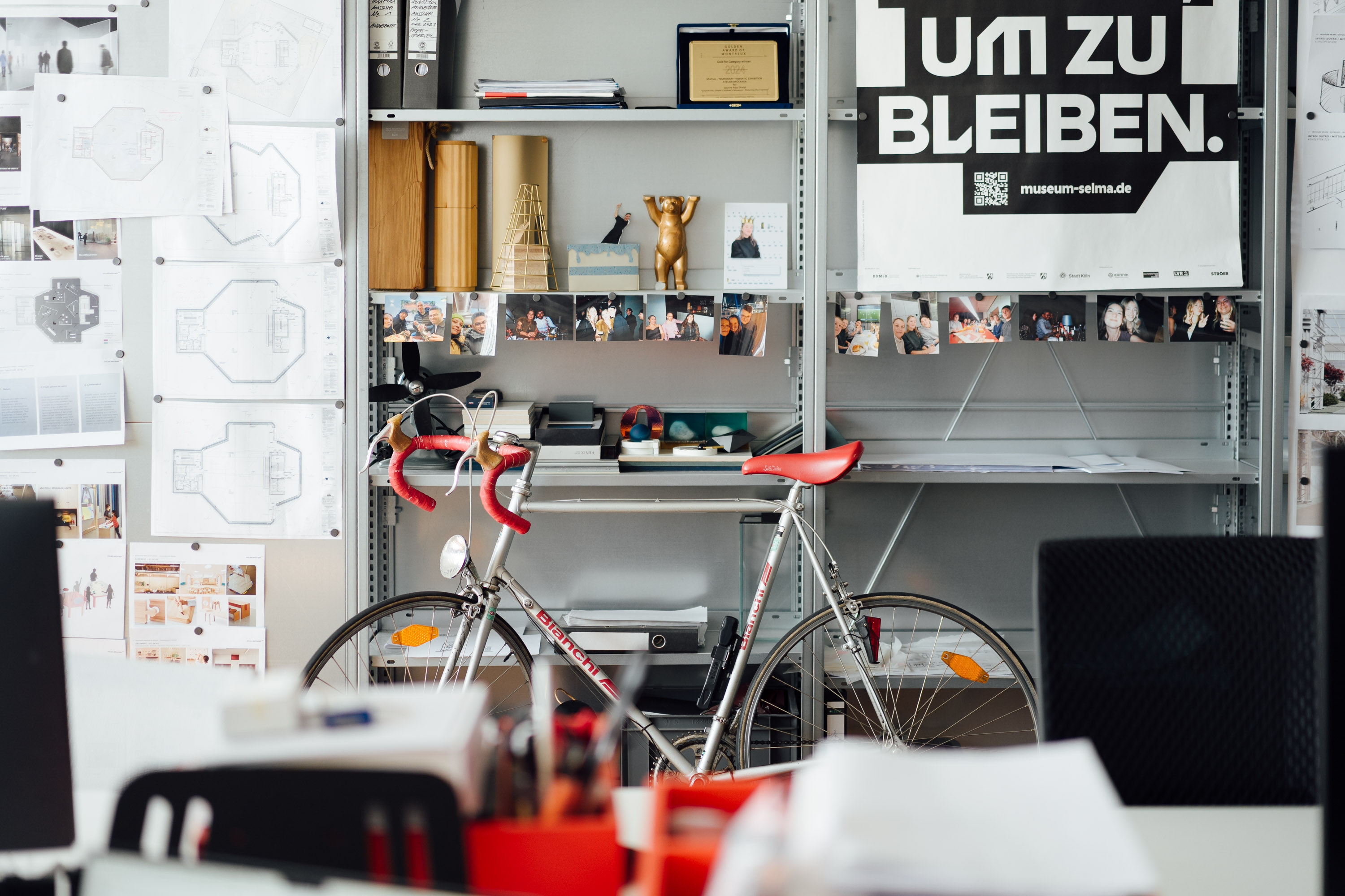 View into a design studio: a silver road bike stands in front of open metal shelving filled with architectural drawings, design materials, and photographs. A large black-and-white poster reads “UM ZU BLEIBEN.” Blurred desks and office chairs are visible in the foreground.