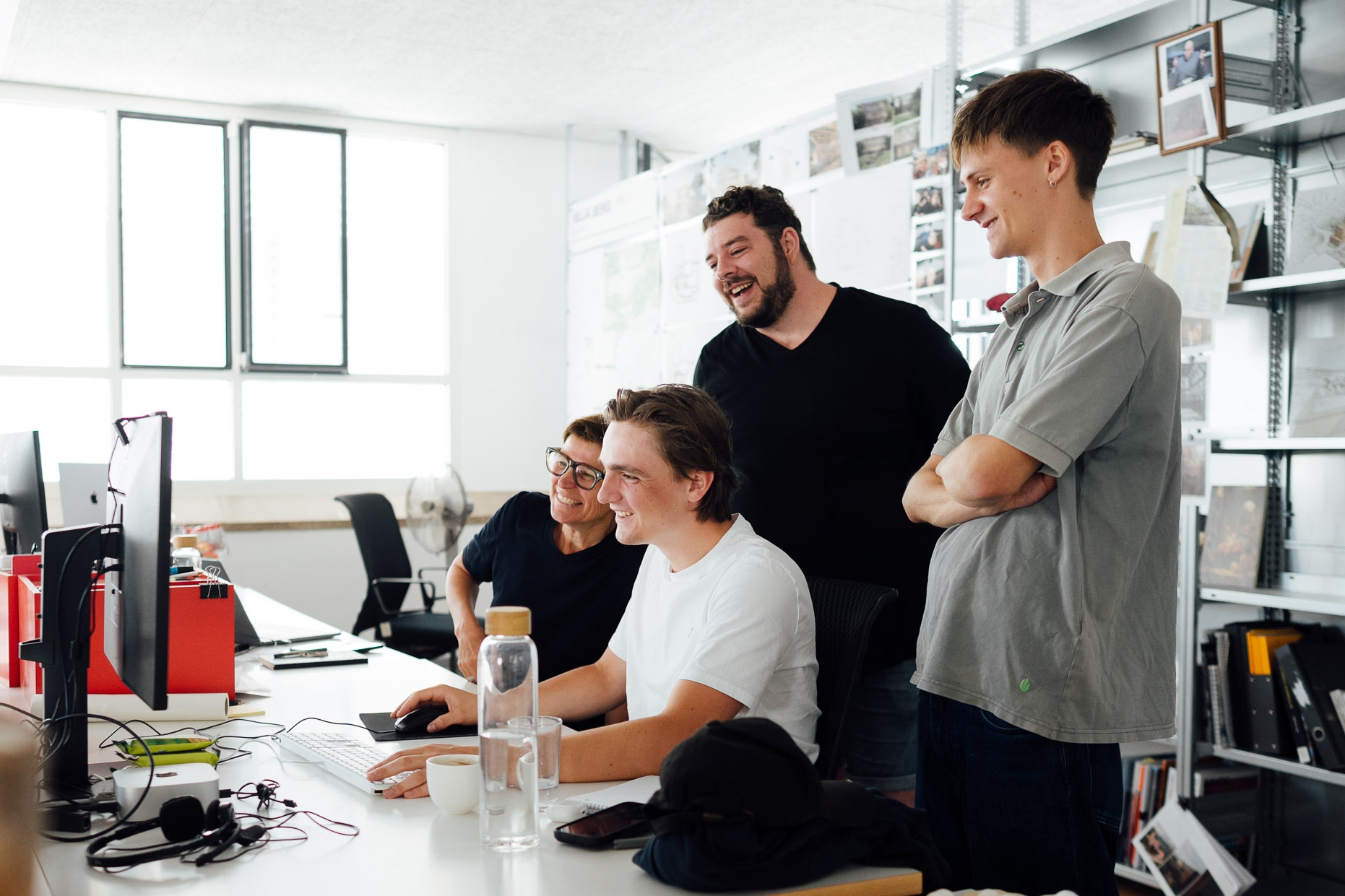 Four colleagues gather around a computer in a bright design studio, smiling as they look at the screen. Architectural plans and project materials are displayed on the walls and shelves around them.