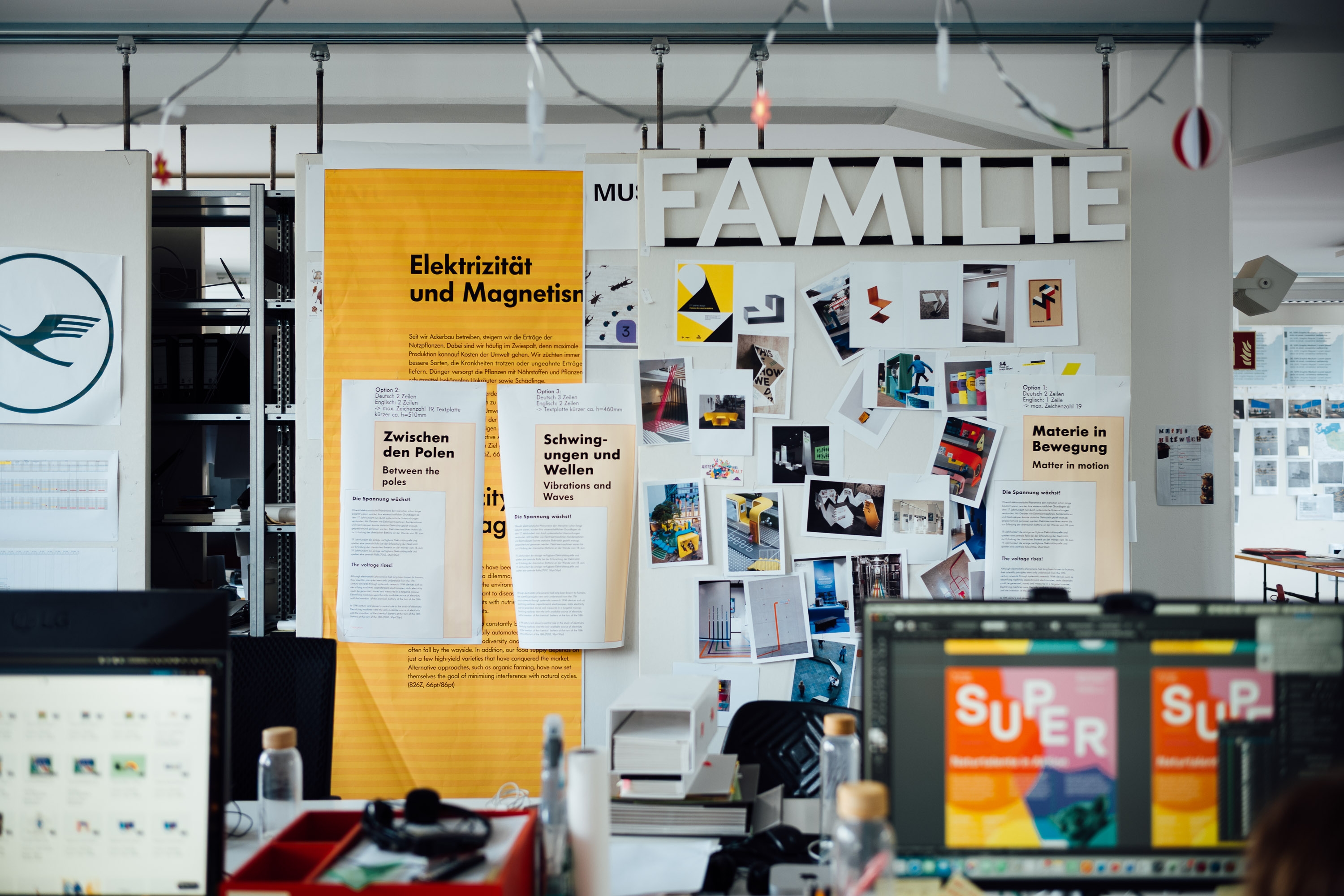 Bürowand mit Ausstellungsentwürfen zu ‚Elektrizität und Magnetismus‘ und ‚Familie‘, gepinnt mit Fotos, Text-Layouts und Moodboard-Bildern über zwei Schreibtischen mit Bildschirmen.
