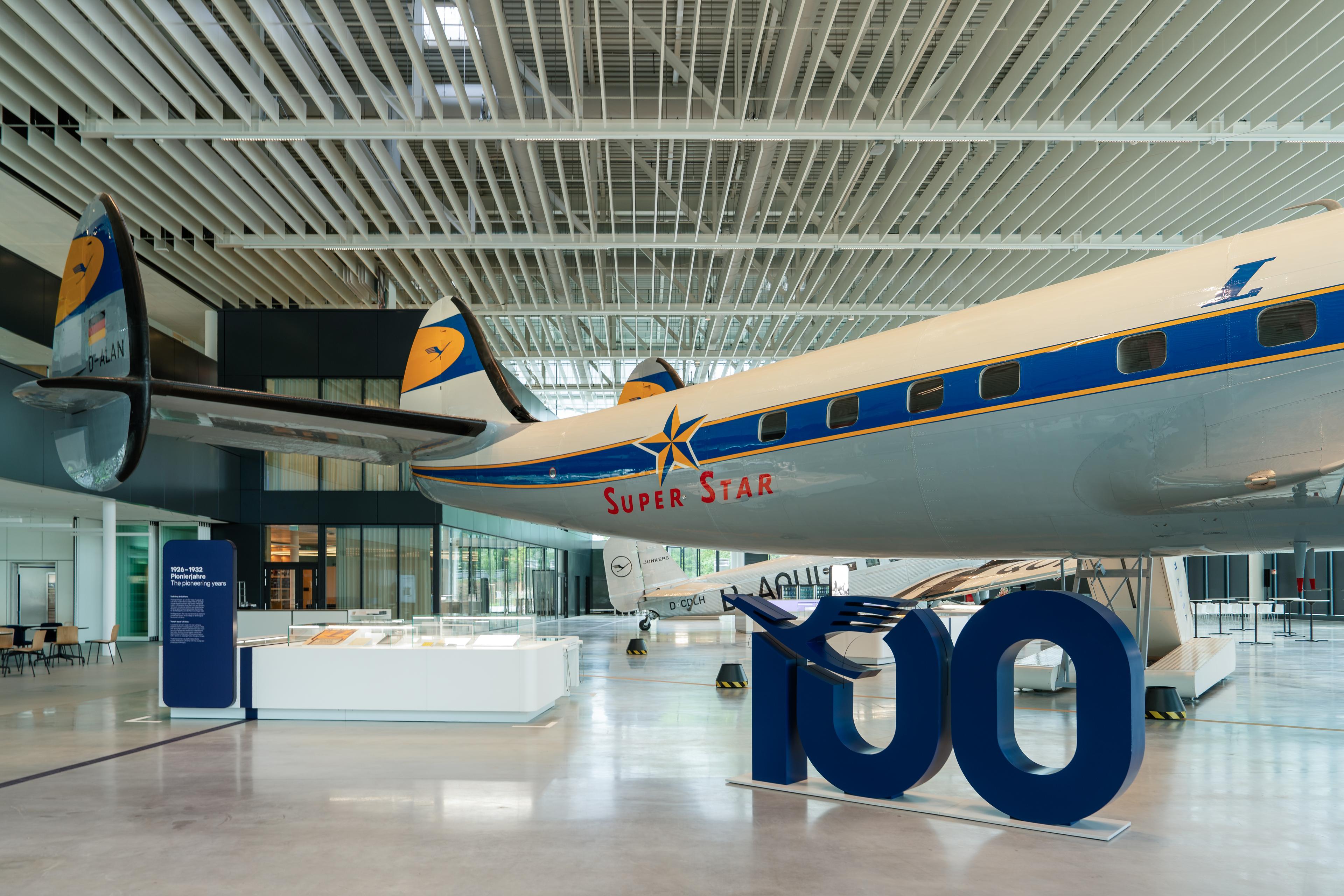 Historic Lufthansa aircraft “Super Star” displayed inside a spacious, light-filled exhibition hall with a high grid ceiling. In the foreground, a large blue “100” sculpture stands next to display cases and information panels about aviation history.