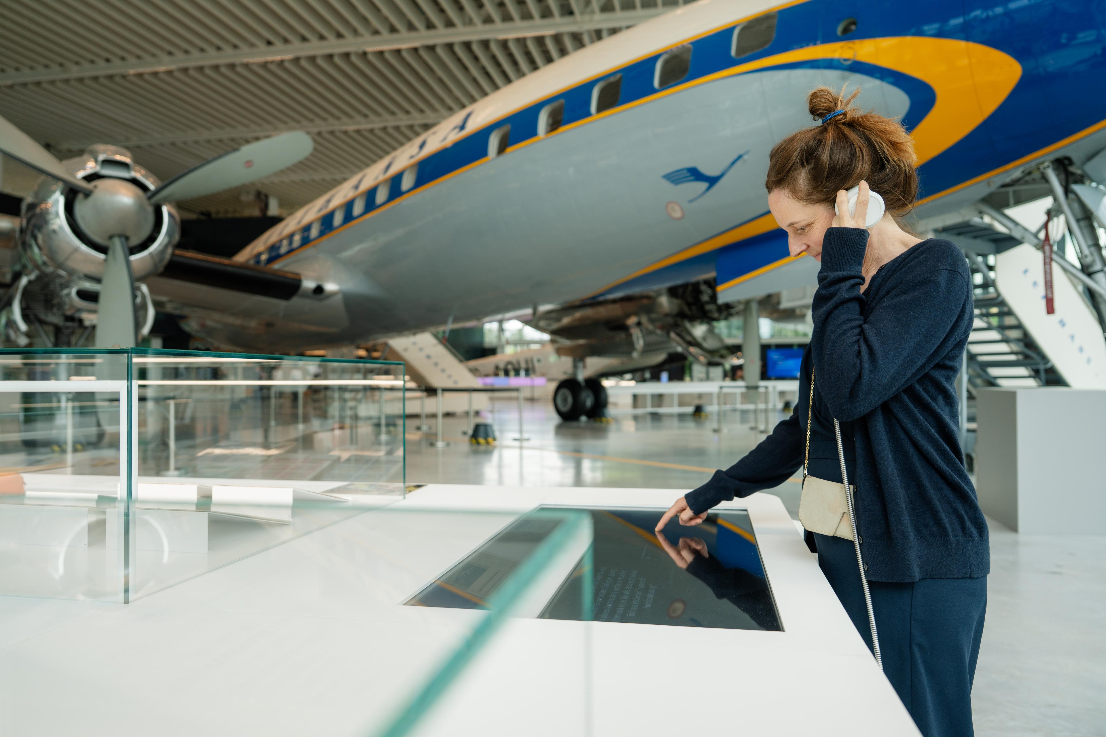 Visitor using an interactive media station inside an aviation exhibition hall, standing in front of a historic Lufthansa aircraft. The spacious interior features display cases, aircraft details, and a high industrial-style ceiling