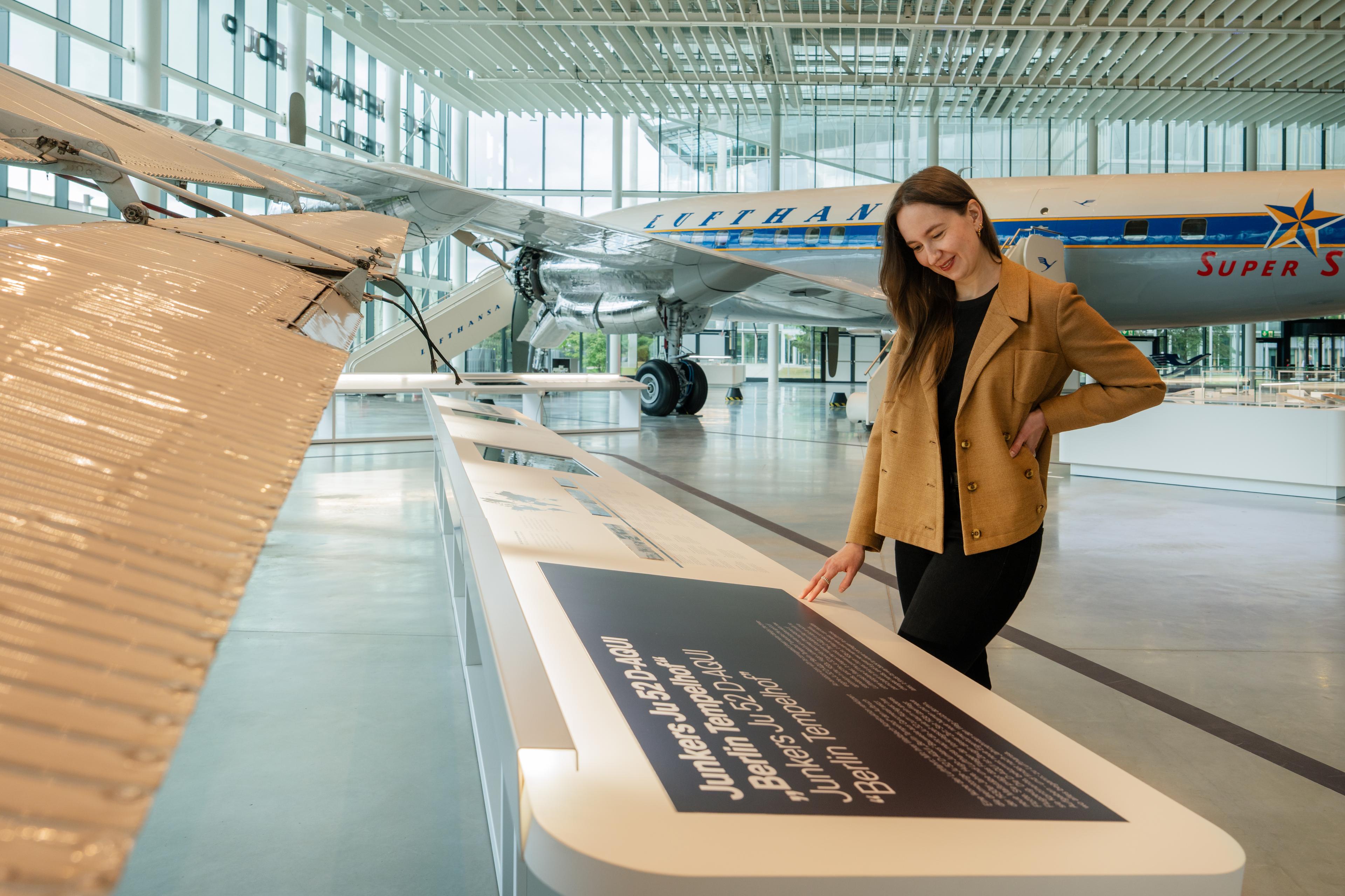 A visitor in a brown jacket reads an exhibition panel inside a spacious aviation exhibition hall with floor-to-ceiling glass façades and a high white grid ceiling. Beside her, the wing and polished metal details of a historic aircraft extend into the foreground, while a restored Lufthansa “Super Star” airplane is displayed in the background among interactive exhibits and open gallery spaces.