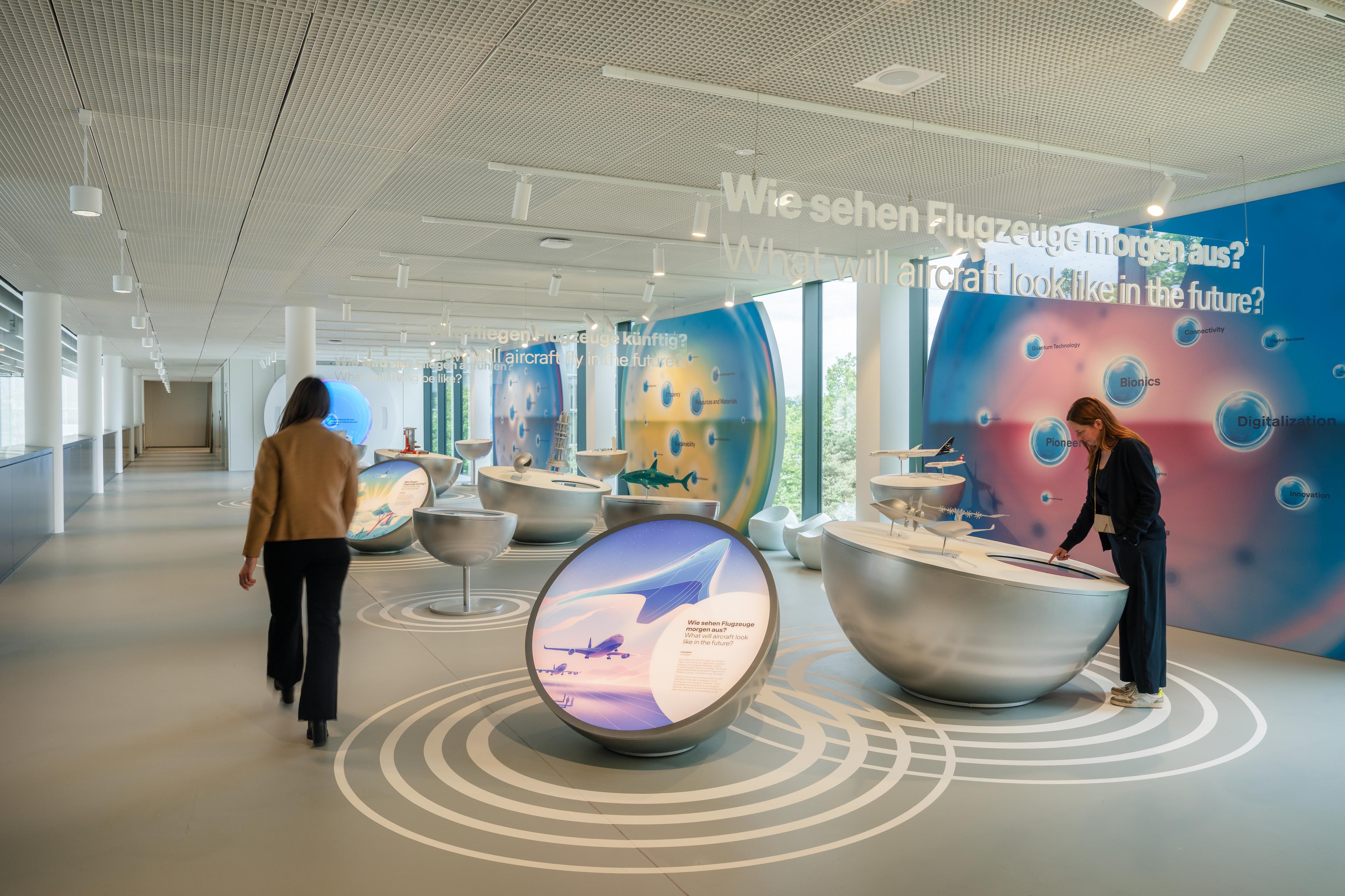 Two visitors explore a futuristic aviation exhibition inside a bright, open gallery space with floor-to-ceiling windows and a perforated ceiling. Curved silver interactive stations and illuminated display tables present concepts for future aircraft technologies, surrounded by large colorful wall graphics and suspended bilingual text reading “What will aircraft look like in the future?”. Circular floor graphics guide visitors through the exhibition environment.