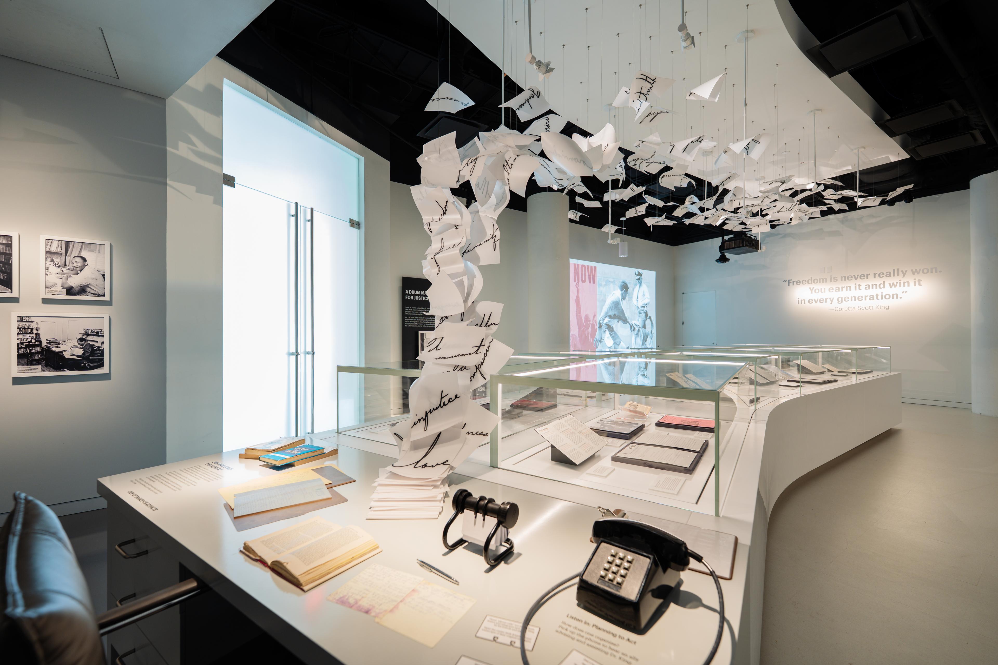 A museum gallery featuring a reconstructed version of Martin Luther King Jr.’s office and writing desk. Books, notes, and office objects are arranged on the desk, while suspended sheets of paper form a dramatic ceiling installation above. Glass cases display archival documents, historic photographs line the walls, and a quotation by Coretta Scott King is visible in the background.