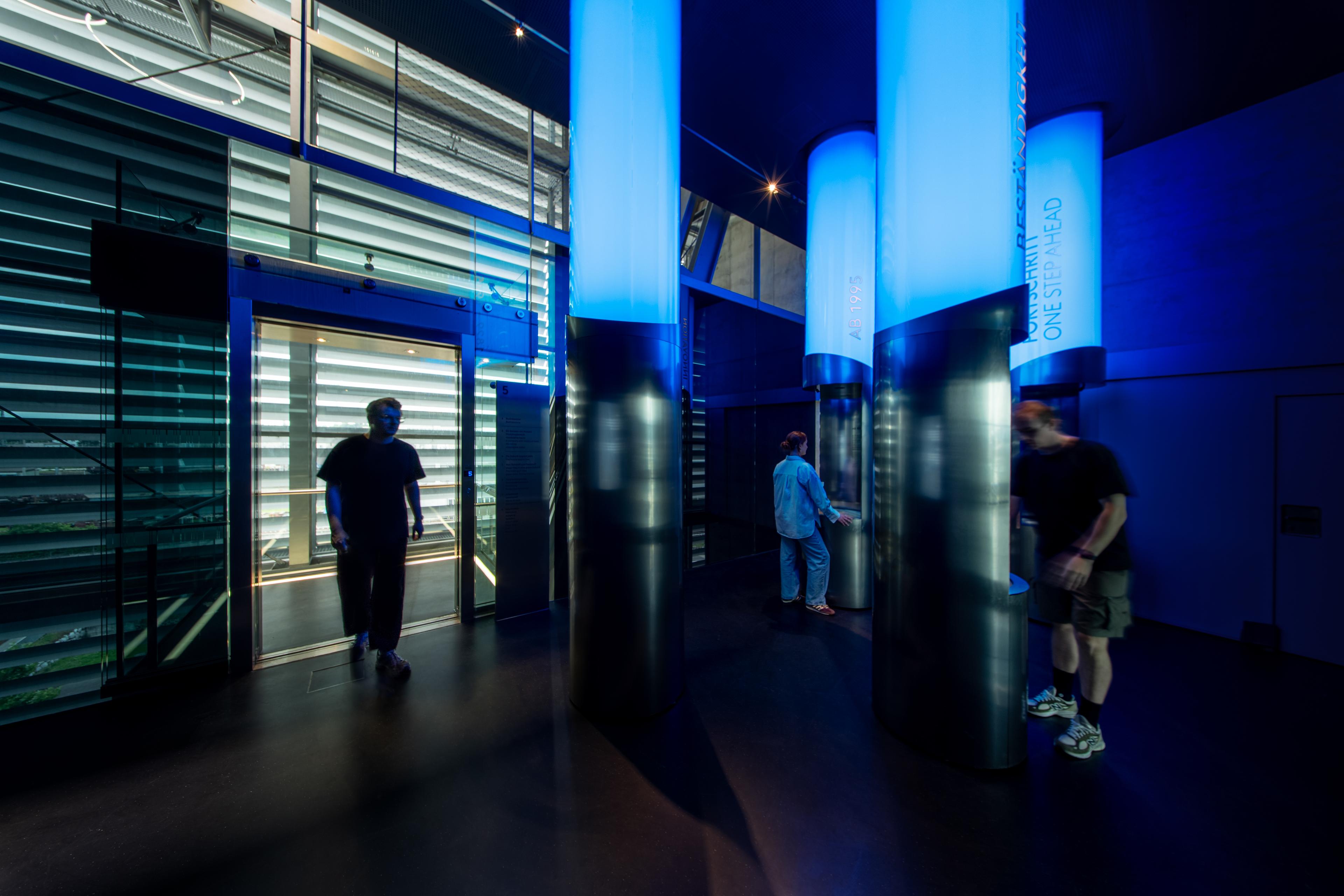 Visitors explore illuminated timeline columns that present key milestones in voestalpine’s history within a dark, immersive exhibition environment
