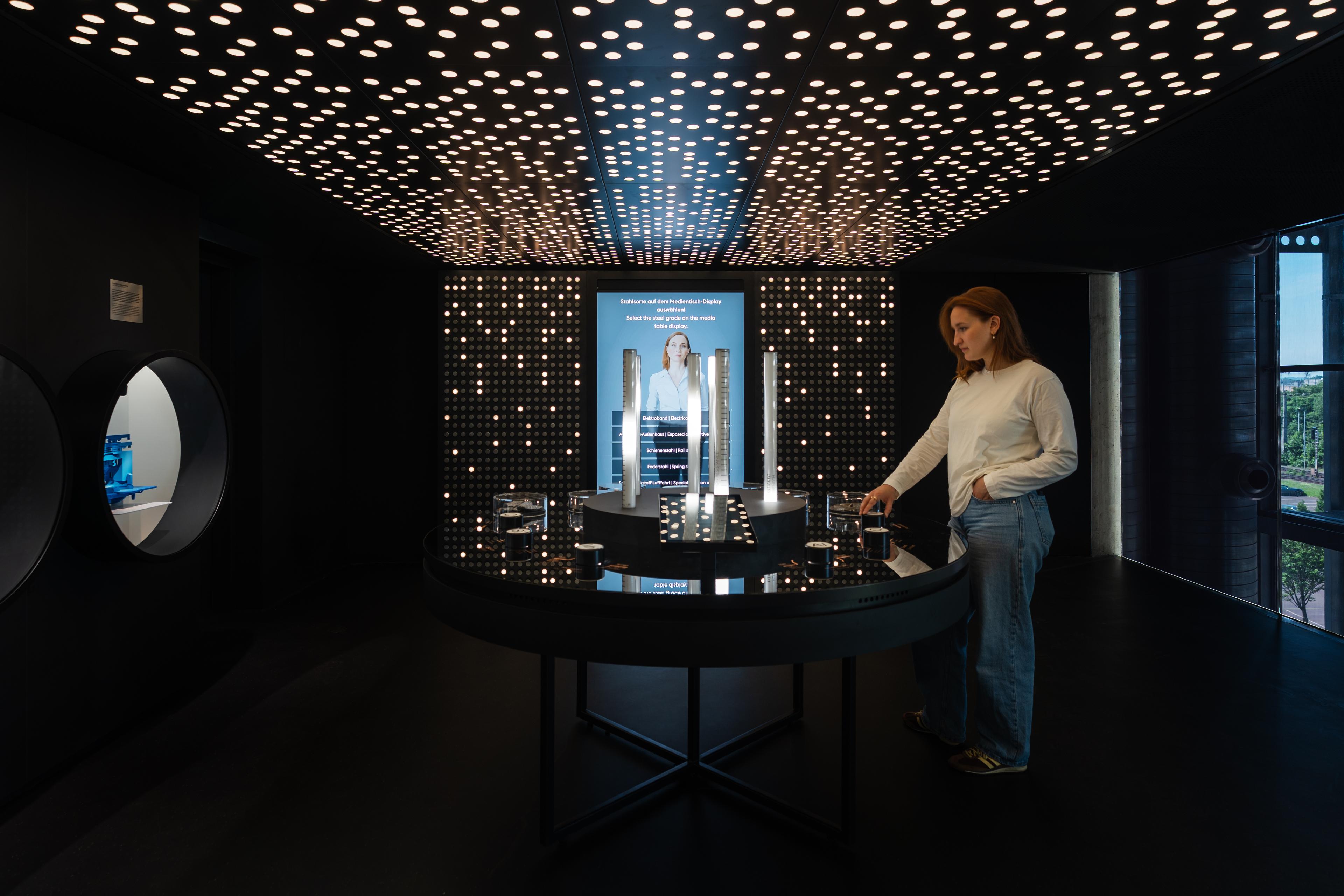 Visitor interacts with a multimedia exhibit on steel grades and material properties, featuring illuminated samples and an immersive light installation inspired by steel production processes