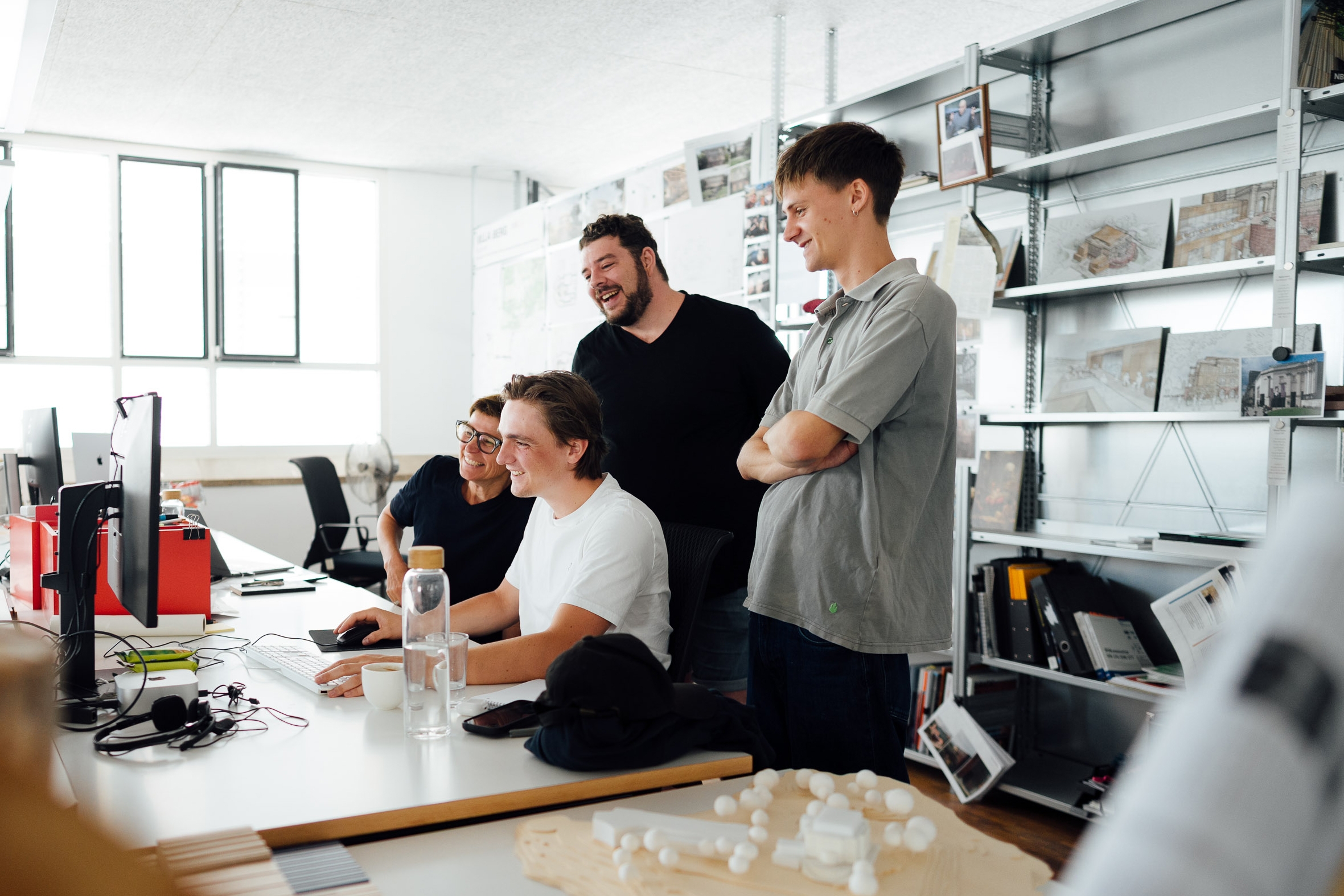 Vier Mitarbeitende stehen und sitzen gemeinsam vor einem Computerbildschirm in unserem hellen Architekturbüro und betrachten lächelnd den Bildschirm. Im Hintergrund sind offene Regale mit Modellen, Plänen, Skizzen und Projektmaterialien zu sehen, im Vordergrund stehen eine Wasserflasche, eine Kaffeetasse und ein Architekturmodell auf dem Tisch. Die Szene vermittelt einen Moment gemeinsamer Zusammenarbeit und Besprechung im Team.