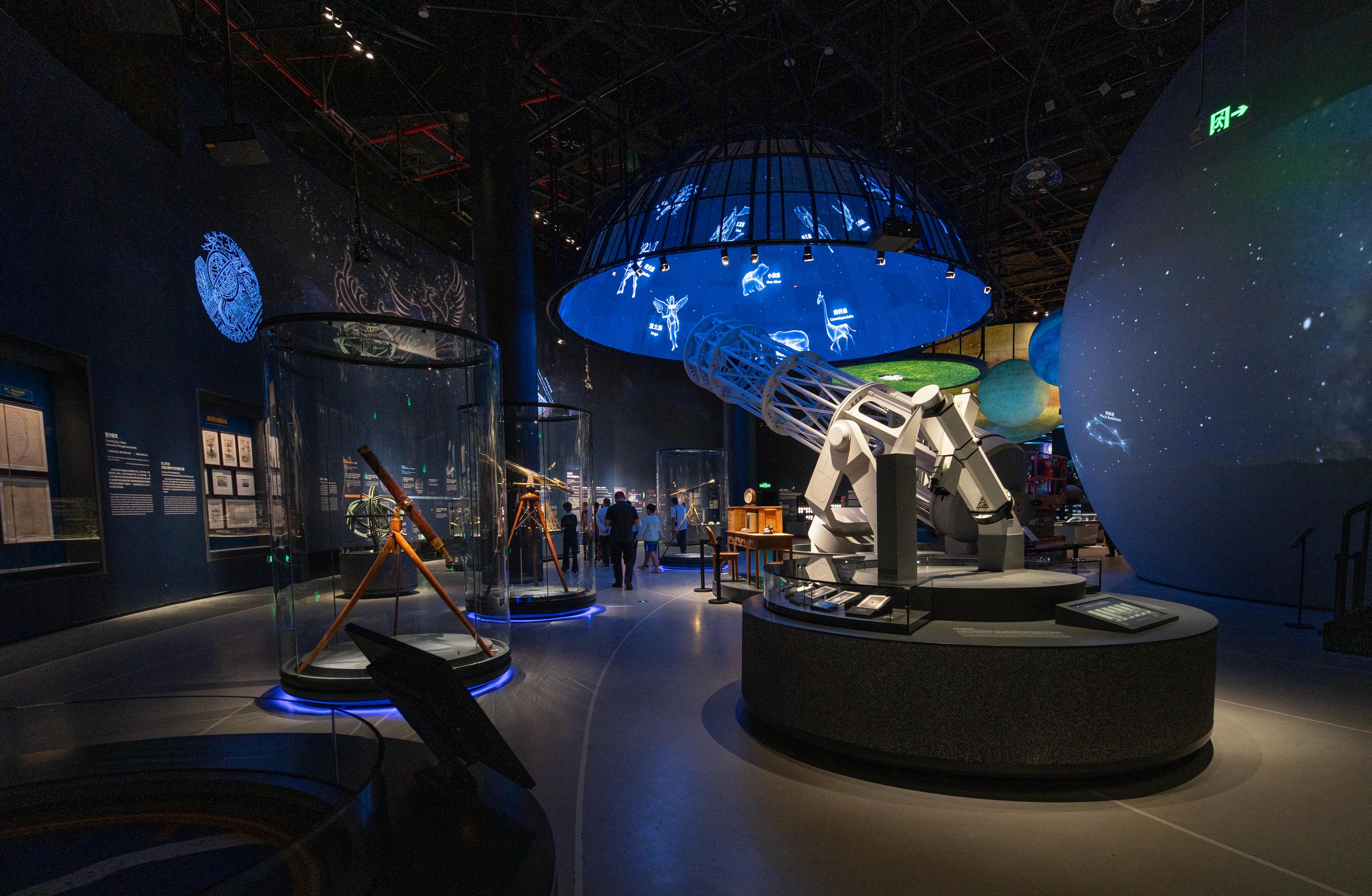 View into the Universe Hall at the Shenzhen Natural History Museum. Historic telescopes and astronomical instruments are displayed in circular glass showcases, while a large telescope model stands prominently in the foreground. Above, a suspended circular media installation projects animated constellations and celestial imagery. Large spherical models of planets and immersive projections create a dark, atmospheric environment that explores humanity’s understanding of the universe.