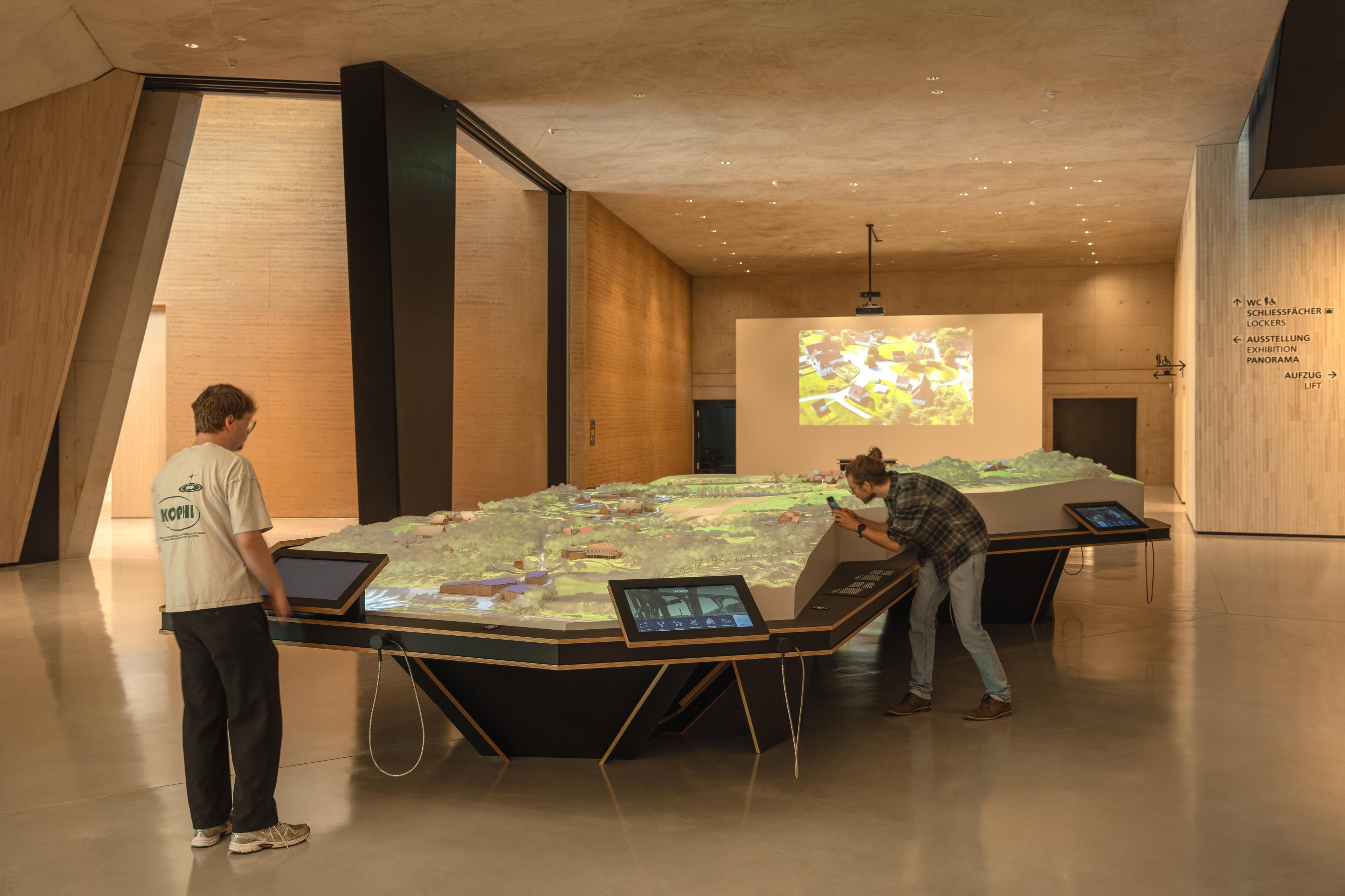Two visitors explore the large scale model of the LWL Museum Hellerlecht, animated by digital projections. Interactive touchscreens surround the model, while a projection on the wall shows an aerial view of the museum grounds.