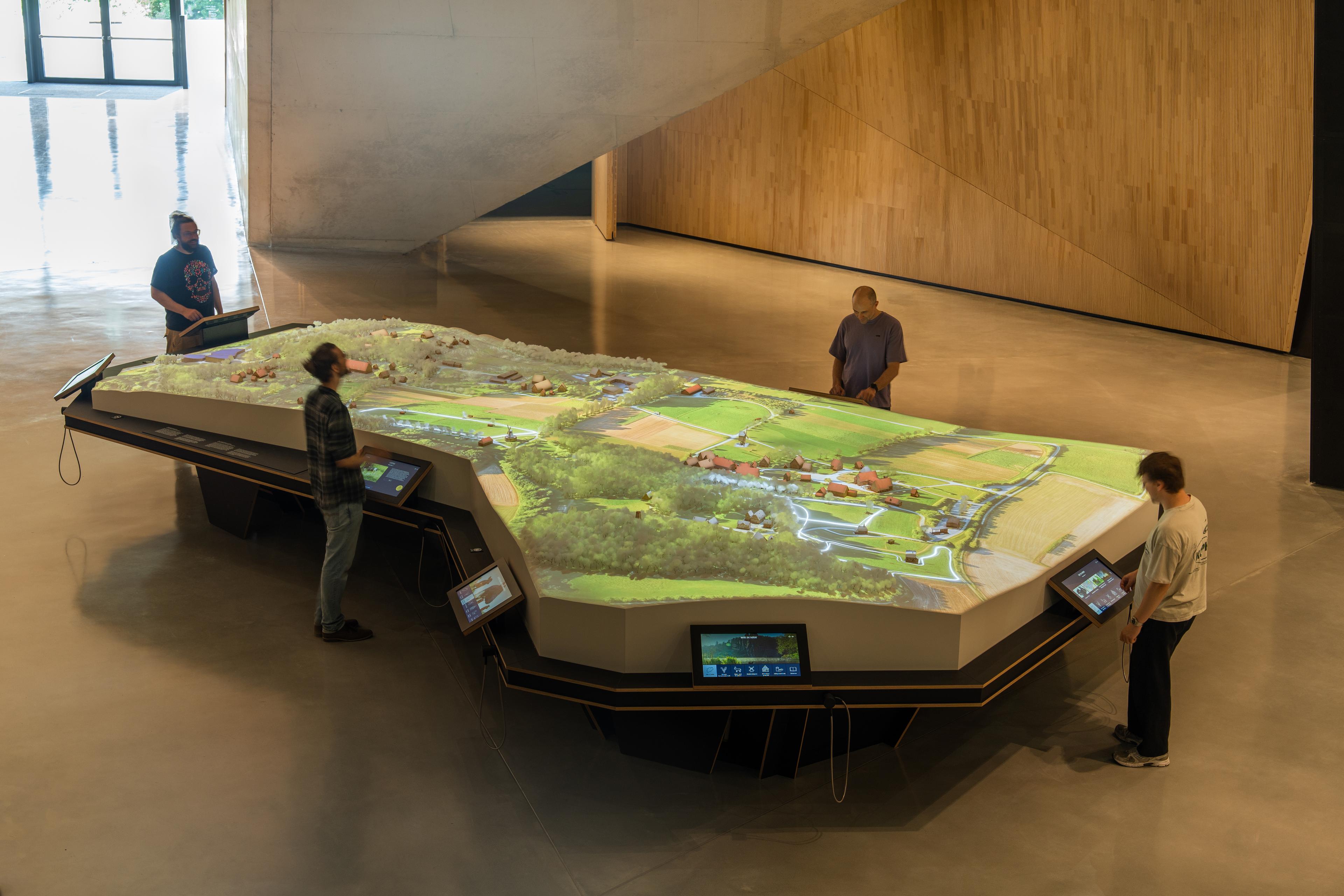 Four visitors interact with a large, illuminated scale model of the LWL Museum Hellerlecht grounds. Digital projections animate the landscape, while touchscreens positioned around the model provide access to information about the open-air museum.