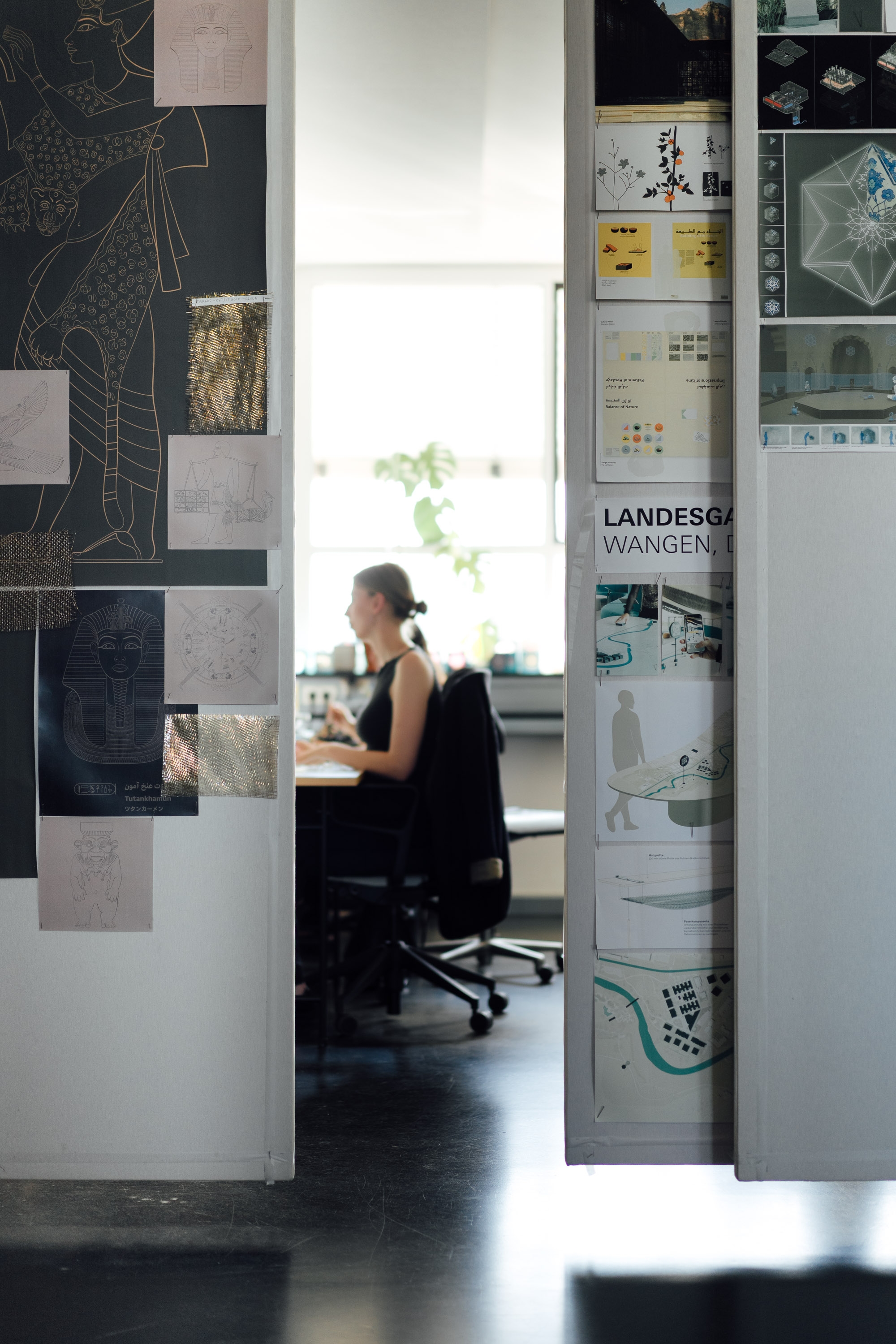 Blick zwischen Pinnwänden mit Entwurfsskizzen hindurch auf eine Person, die im Hintergrund an einem Schreibtisch arbeitet.