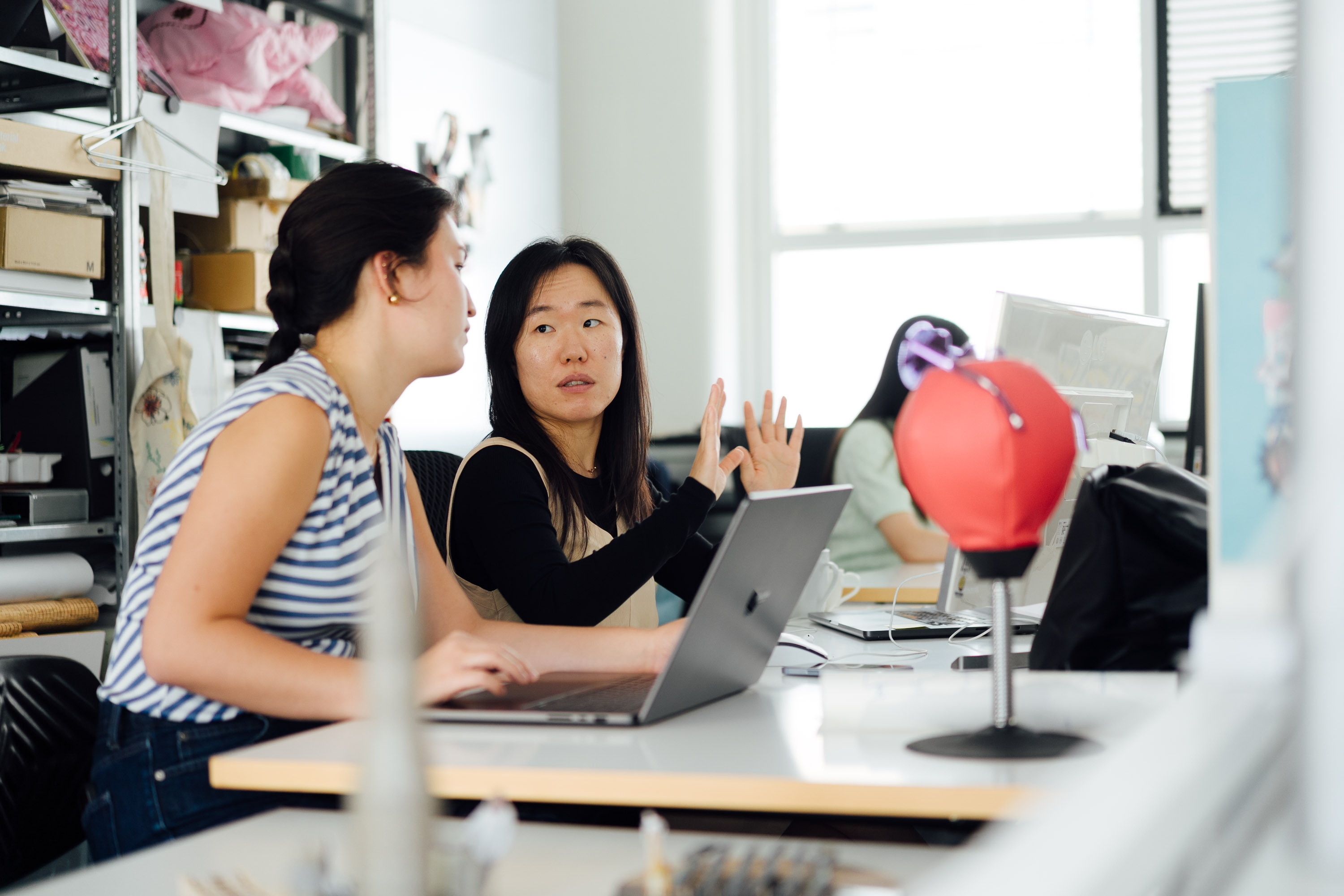 Zwei Frauen sitzen an einem Tisch in einem hellen Büro und unterhalten sich. Eine von ihnen gestikuliert neben einem geöffneten Laptop. Im Hintergrund arbeitet eine weitere Person an einem Computer; links stehen Regale mit Ordnern und Materialien.