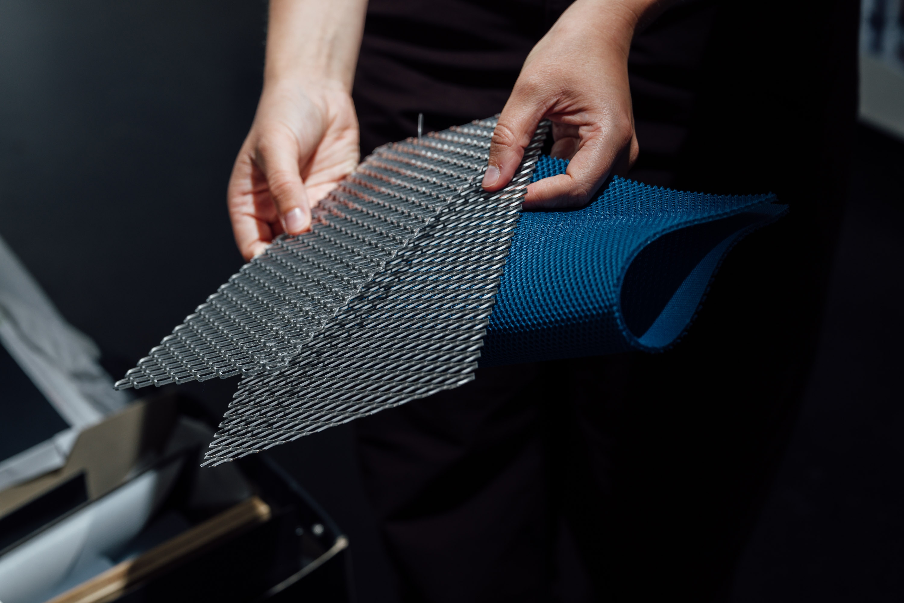 Nahaufnahme von Händen, die mehrere übereinanderliegende Materialproben aus Metallgewebe und blauem Kunststoff halten. Die Proben haben unterschiedliche Strukturen und Maschenweiten.